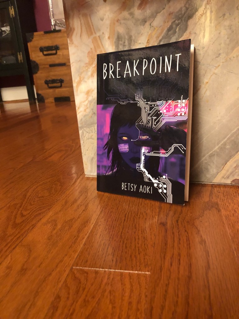Picture of  the poetry collection Breakpoint leaning against a tan marble coffee table base on a rust-colored wooden floor. Furniture in the background.  Breakpoint cover features a dark haired woman making a peace sign as white silicon circuitry flows around her face.